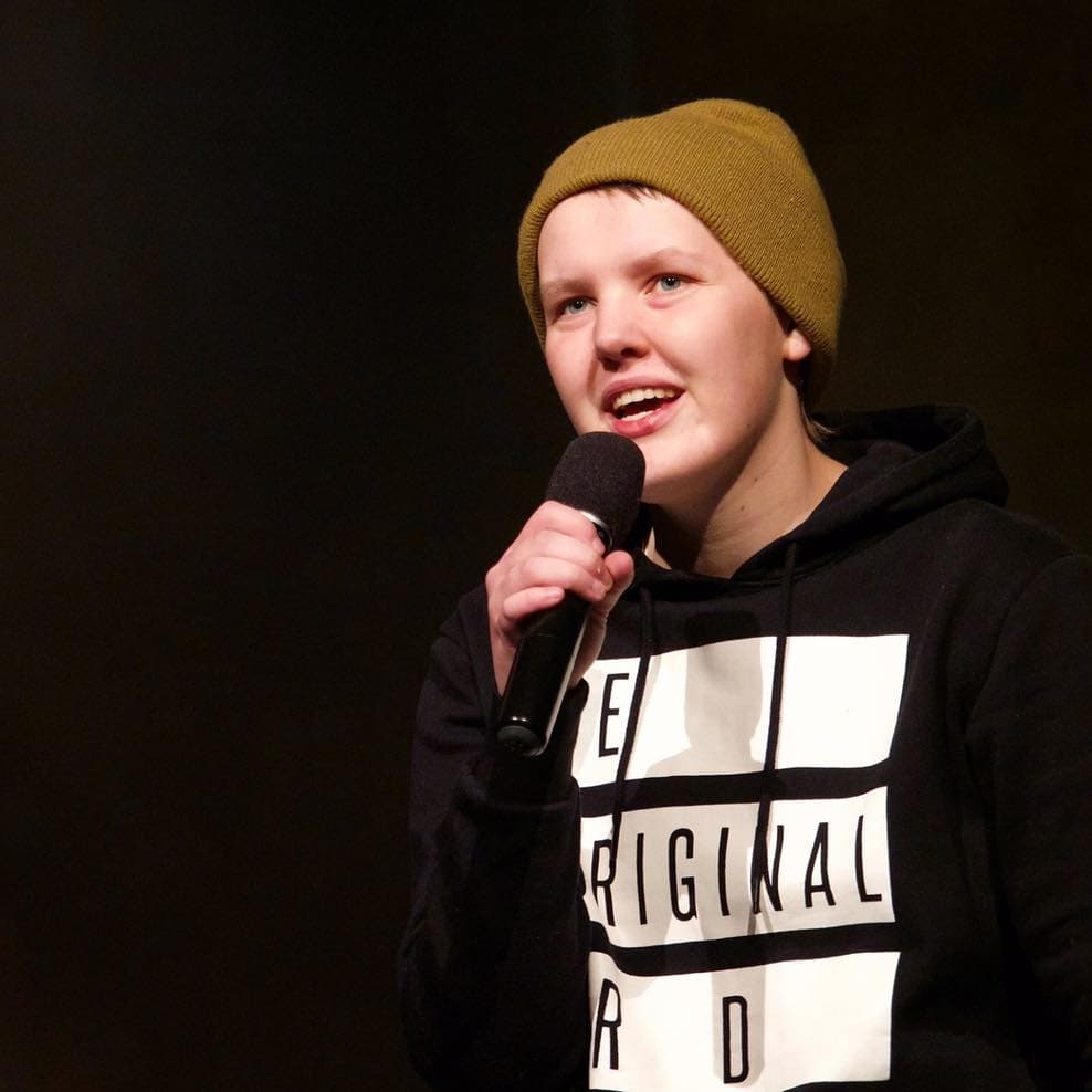 Young person in a yellow beanie and black hoodie speaks into a microphone on stage.