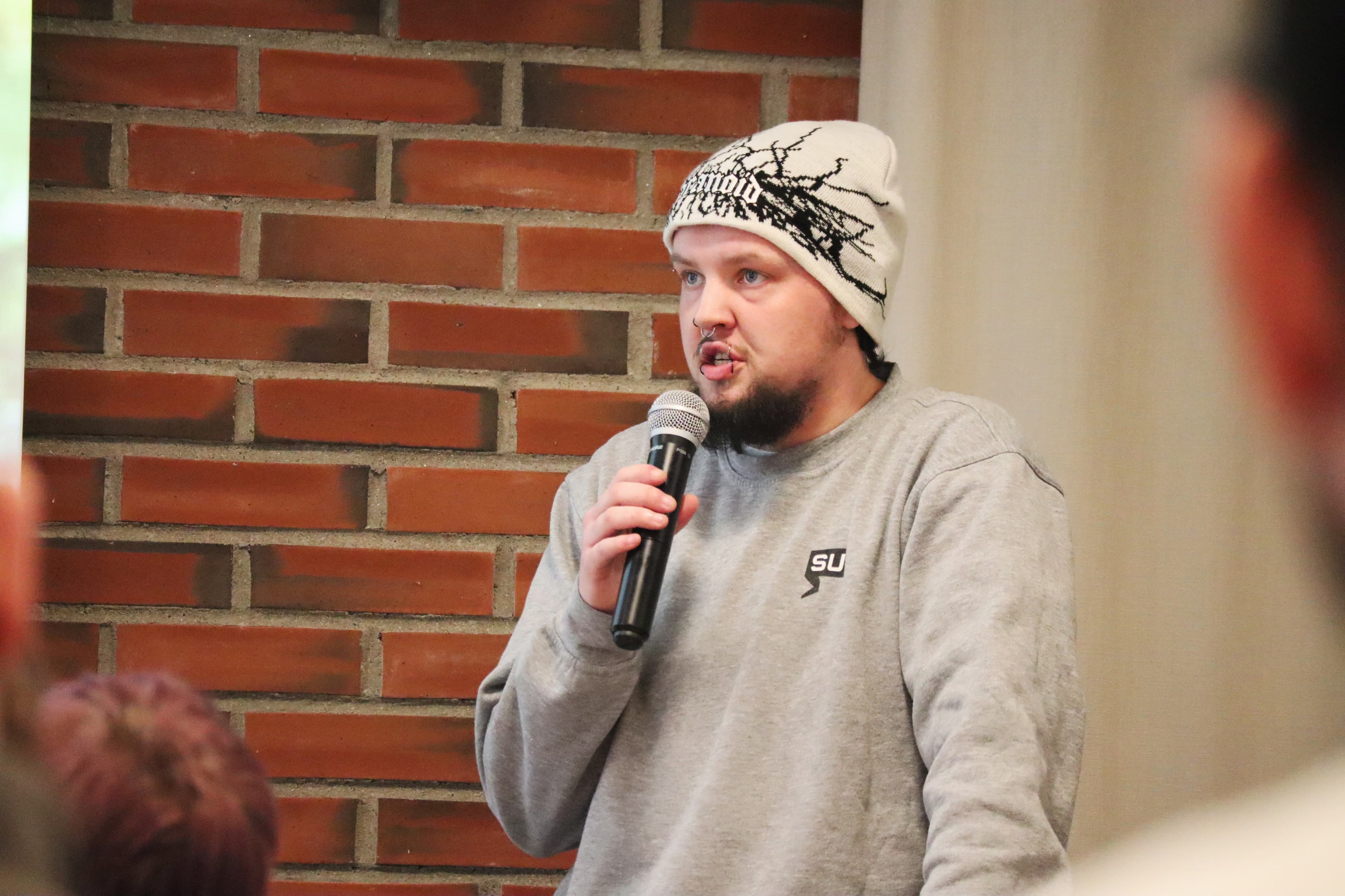 Bearded man in a white beanie speaking into a microphone against a brick wall.