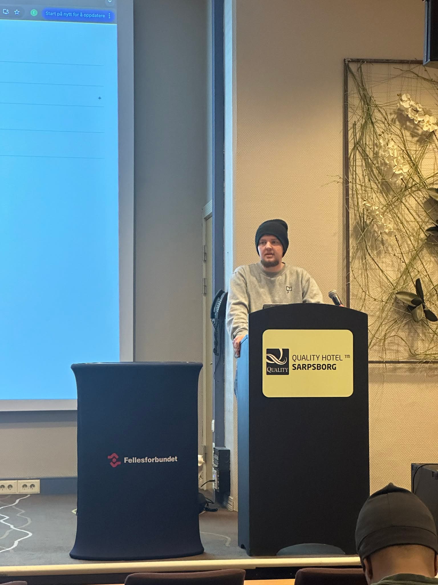 Man in a beanie speaks at a Quality Hotel Sarpsborg podium during a presentation.