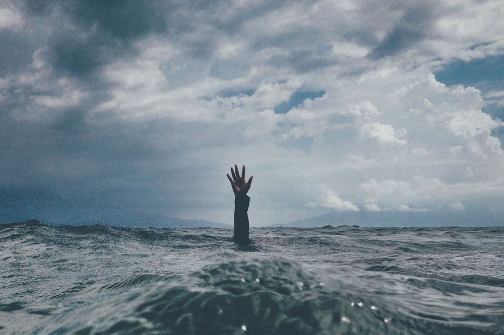 A hand reaches out from choppy ocean water under a vast, dark and dramatic sky.
