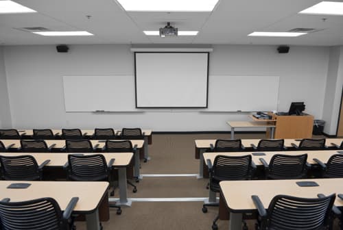 Empty lecture hall with rows of desks, black chairs, a projector screen, and whiteboards.