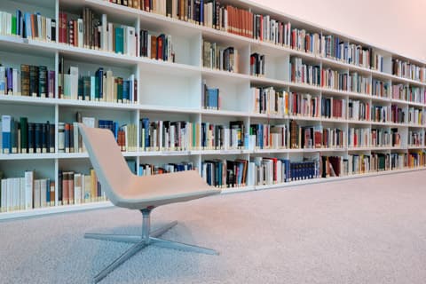 Modern beige chair in front of large white bookshelves filled with many colorful books.