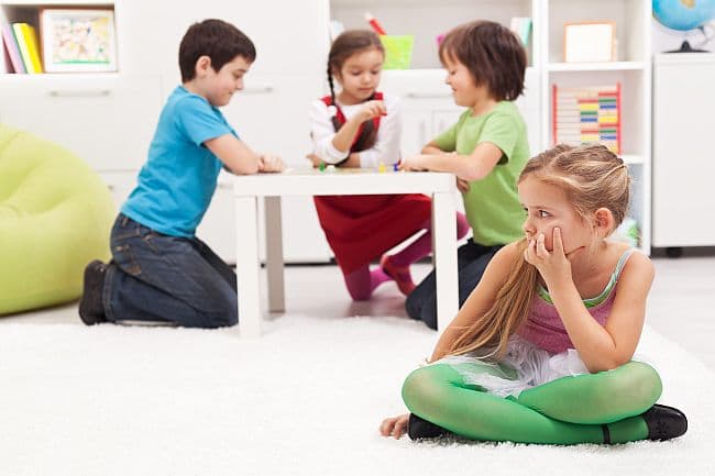 Sad girl sits alone on the floor while other children play together behind her.