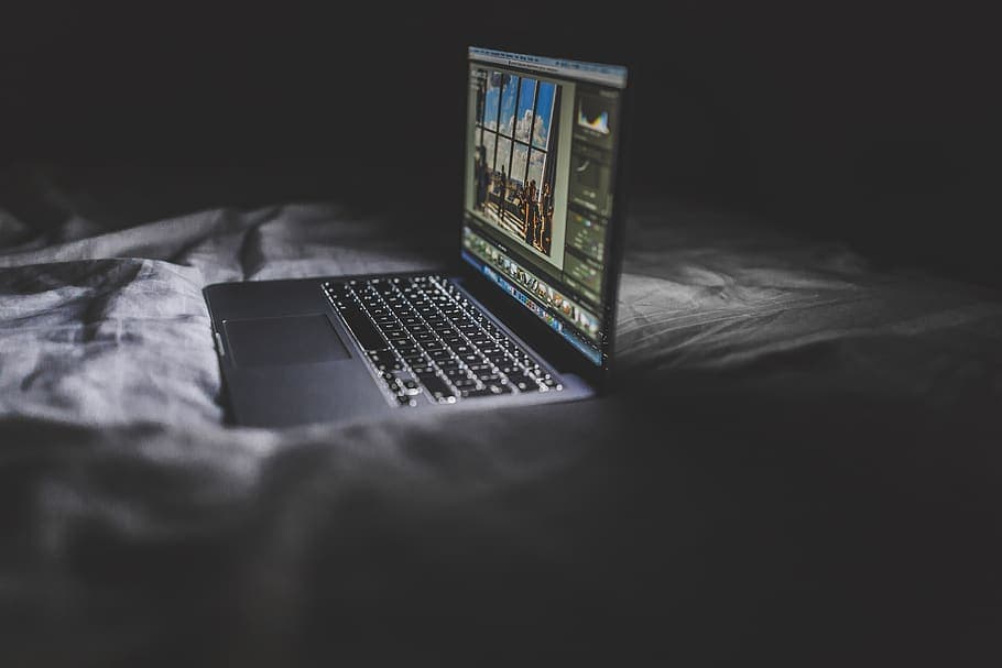 Open laptop on a rumpled bed in a dark room, displaying photo editing software.